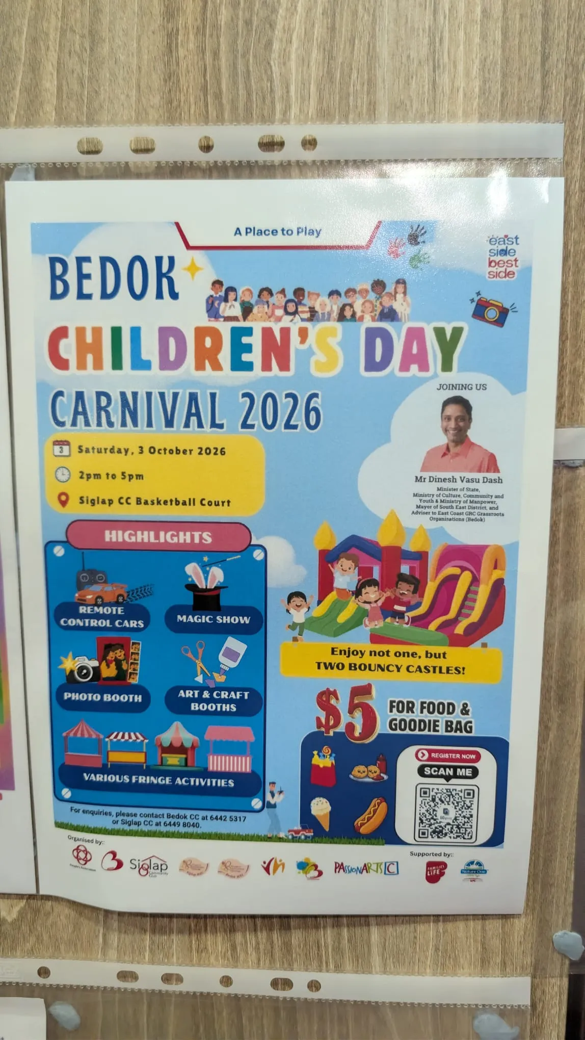 A printed flyer in a plastic sleeve on a wooden wall: Bedok Children's Day Carnival 2026, Saturday 3 October, 2pm to 5pm, Siglap CC basketball court, with bouncy castles, a magic show and a $5 food and goodie bag
